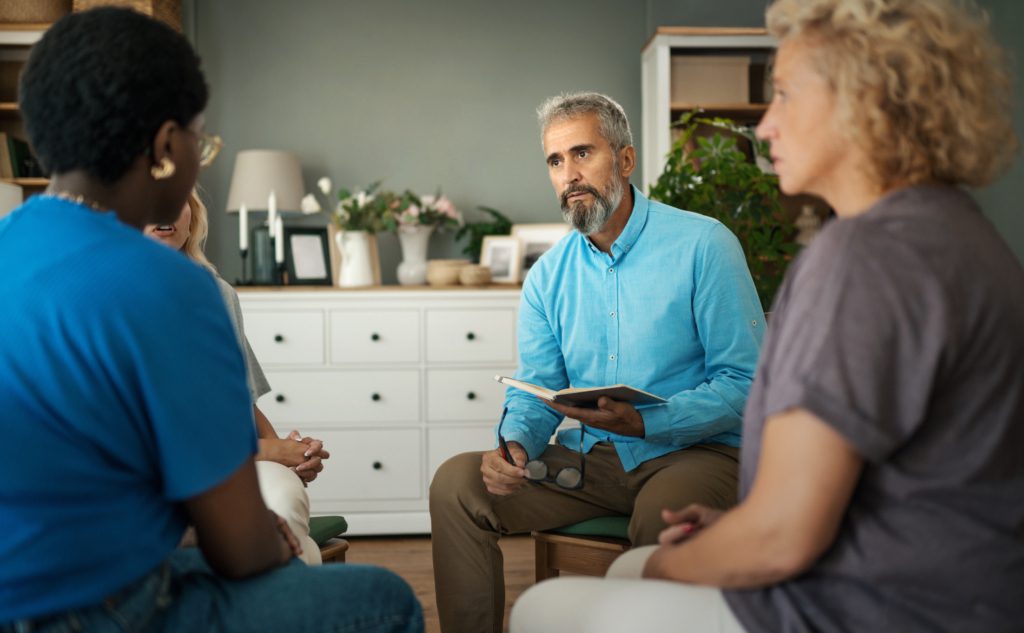 An older male psychiatrist leads a group therapy session in a modern home-like environment